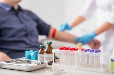 pacjent oddaje krew, przed nim na stole leżą probówki w próbkami krwi i odczynniki laboratoryjne