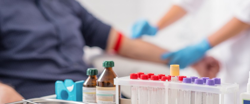 pacjent oddaje krew, przed nim na stole leżą probówki w próbkami krwi i odczynniki laboratoryjne