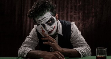 Mężczyzna w stroju Jokera siedzi przy stole w ciemmności i pokazuje kartę Joker