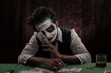 Mężczyzna w stroju Jokera siedzi przy stole w ciemmności i pokazuje kartę Joker