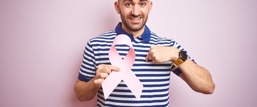 Młody mężczyzna trzyma różową wstążkę raka piersi ze zdziwioną twarzą, wskazuje palcem na siebie