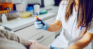 asystent laboratoryjny w mundurze, w masce i gumowych rękawiczkach trzymający probówkę z krwią, podczas gdy pacjent siedzi i trzyma absorbent