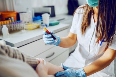 asystent laboratoryjny w mundurze, w masce i gumowych rękawiczkach trzymający probówkę z krwią, podczas gdy pacjent siedzi i trzyma absorbent