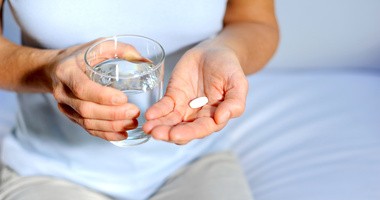 Kobieta trzyma w jednej dłoni tabletkę przeciwbólową, a w drugiej szklankę wody do popicia