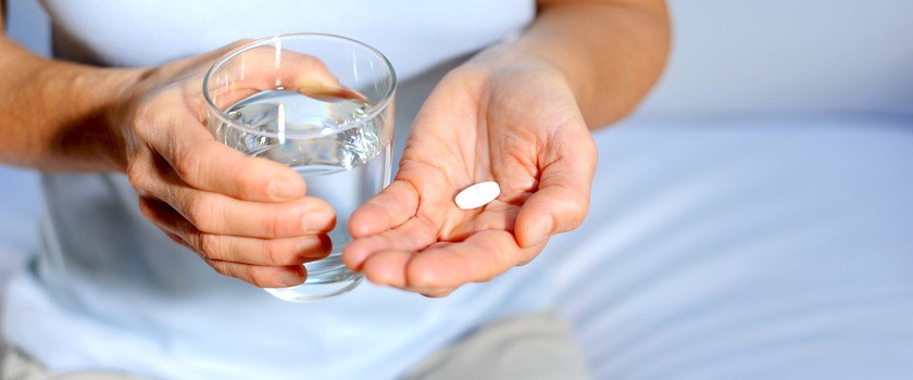 Kobieta trzyma w jednej dłoni tabletkę przeciwbólową, a w drugiej szklankę wody do popicia