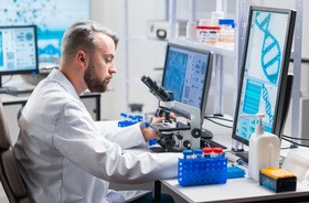 Naukowiec siedzi przy mikroskopie i przed monitorem, na którym wyświetla się podwójna nić DNA
