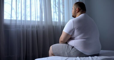 Mężczyzna z otyłością siedzi smutny na brzegu łóżka w ciemnym pokoju