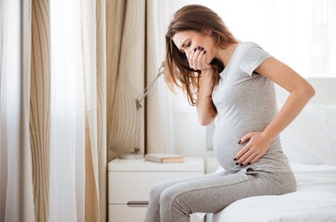 Kobieta w ciąży siedzi na brzegu łóżka, zakrywając sobie usta z powodu mdłości