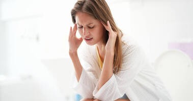 Młoda kobieta siedzi na sedesie z migreną i problemami żoładkowymi