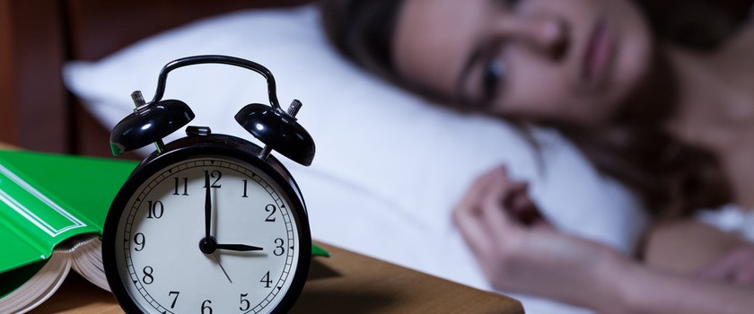 Kobieta wybudzona w nocy leży w łóżku, na pierwszym planie zegarek-budzik wskazujący godzinę 3 w nocy