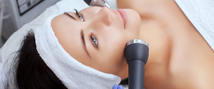 Kosmetolog wykonuje zabieg ultradźwiękowym oczyszczeniem skóry twarzy młodej kobiety w gabinecie kosmetycznym.