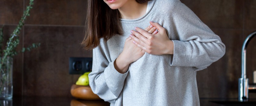 Młoda kobieta w szarym swetrze trzyma się za serce z grymasem na twarzy