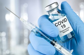 Dłoń w niebieskich rękawiczkach medycznych trzyma fiolkę ze szczepionką na COVID-19 i strzykawkę