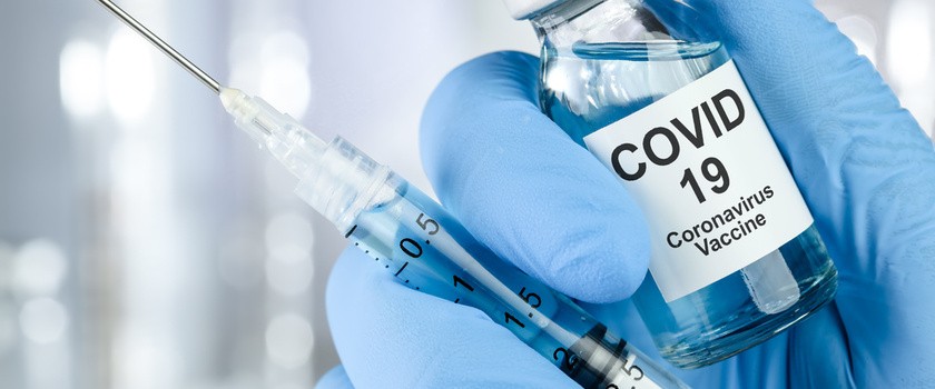 Dłoń w niebieskich rękawiczkach medycznych trzyma fiolkę ze szczepionką na COVID-19 i strzykawkę