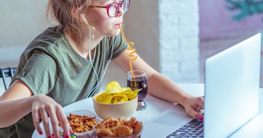 Młoda dziewczyna siedzi przed komputerem, jedząc i pijąc niezdrowe produkty, ultra-przetworzone