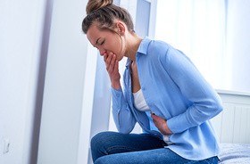 Kobieta ciepriąca z powodu mdłości w grypie jelitowej, siedzi i trzyma się za brzuch, zakrywając sobie także usta dłonią