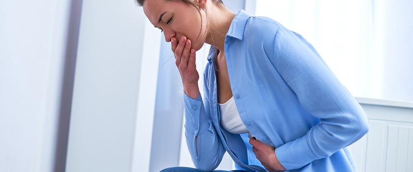 Kobieta ciepriąca z powodu mdłości w grypie jelitowej, siedzi i trzyma się za brzuch, zakrywając sobie także usta dłonią