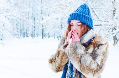 Dziewczyna w zimowych ubraniach stoi na tle zaśniezonego lasu, chuchając w swoje dłonie, które ma zmarznięte