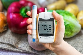 Glukometr i pen z insuliną trzymany w dłoni na tle owoców i warzyw