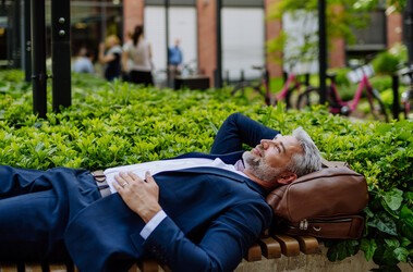 Męzczyzna typu biznesmen próbujący zachować work-life balance - wypoczywa w garniturze na ławce pod korporacją