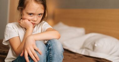 Zmartwiona dziewczynka w wieku ok. 7 lat siedzi na krawędzi łóżka. Moczenie nocne u dzieci, enureza u dzieci.