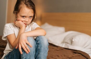 Zmartwiona dziewczynka w wieku ok. 7 lat siedzi na krawędzi łóżka. Moczenie nocne u dzieci, enureza u dzieci.