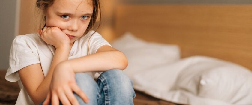 Zmartwiona dziewczynka w wieku ok. 7 lat siedzi na krawędzi łóżka. Moczenie nocne u dzieci, enureza u dzieci.