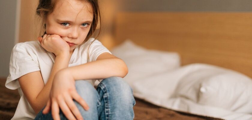 Zmartwiona dziewczynka w wieku ok. 7 lat siedzi na krawędzi łóżka. Moczenie nocne u dzieci, enureza u dzieci.